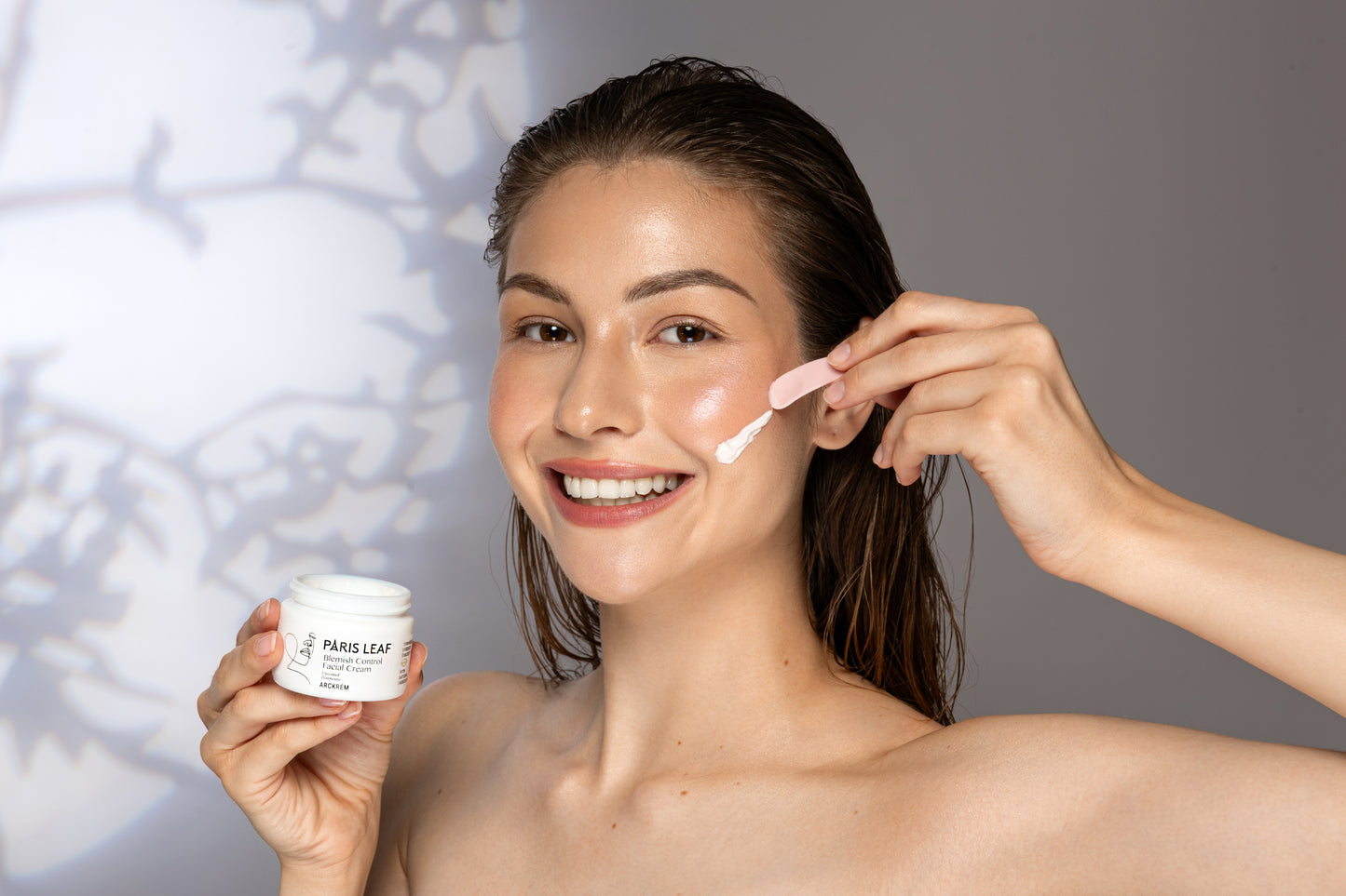 Smiling woman applying Parisleaf Blemish Control Facial Cream to her cheek with a skincare spatula.